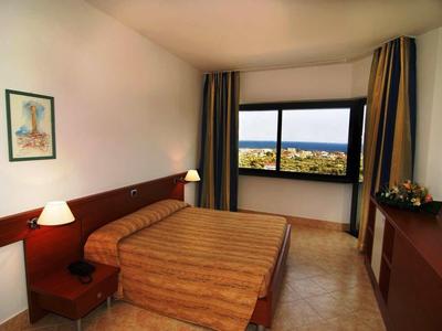 Einfaches Doppelzimmer mit Doppelbett, Nachttischen, Lampe, Fenster mit Meerblick und Vorhängen.