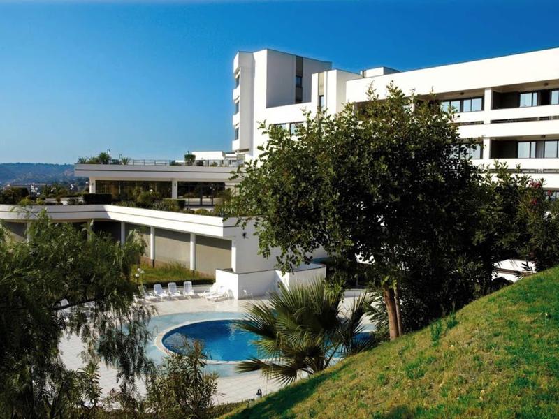 Modernes weißes Hotelgebäude mit Pool und grünem Hang bei klarem blauem Himmel.