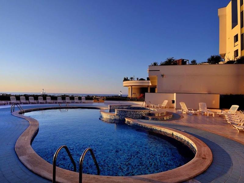 Schwimmbad mit geschwungener Form, umgeben von weißen Liegestühlen vor klarem blauem Himmel.