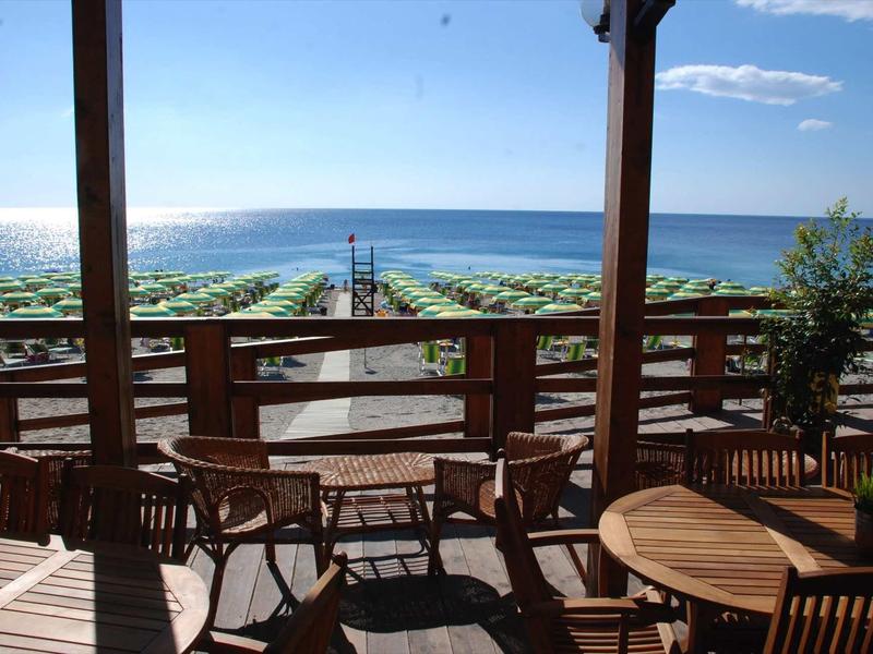 Blick von überdachter Terrasse mit Holzmöbeln auf Strand mit grünen Sonnenschirmen und blauem Meer.