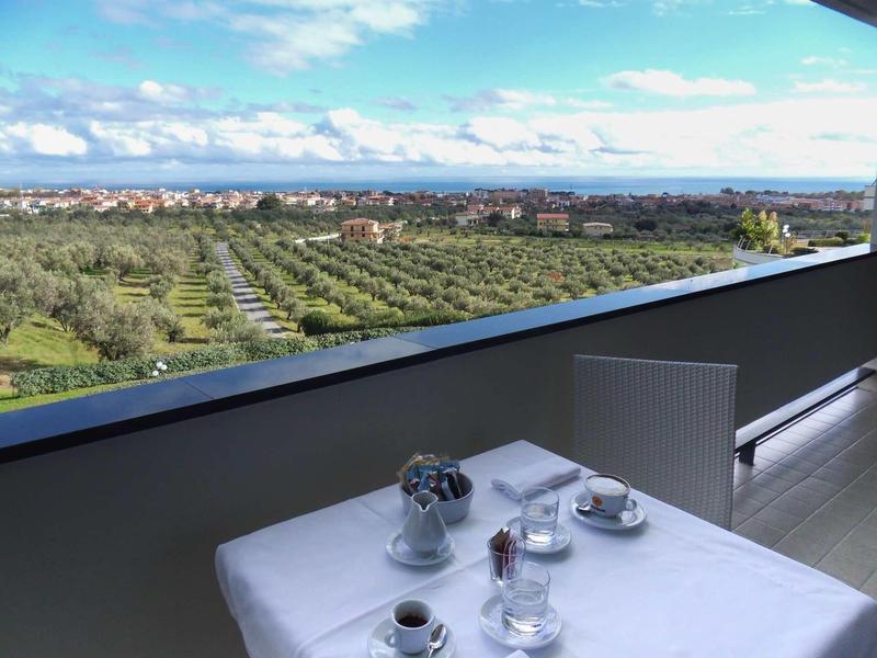 Balkon mit grauem Geländer, weißem Tisch, zwei Tassen und weiter Sicht auf Felder und Stadt.