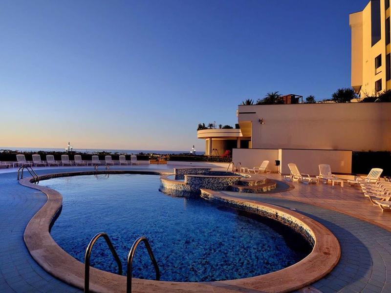 Ein großer, geschwungener Pool mit blauer Wasseroberfläche, umgeben von weißen Liegestühlen bei klarem Himmel.