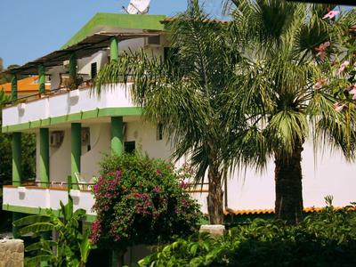 Maison de vacances blanche avec accents verts, balcon, entourée d'une végétation tropicale luxuriante.