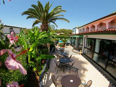 Balkon mit Tischen, Stühlen, Palmen und Blüten, angrenzend an ein Gebäude mit Balkonen.