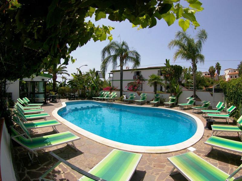 Piscine avec transats verts entourée de palmiers et plantes sous un ciel bleu.