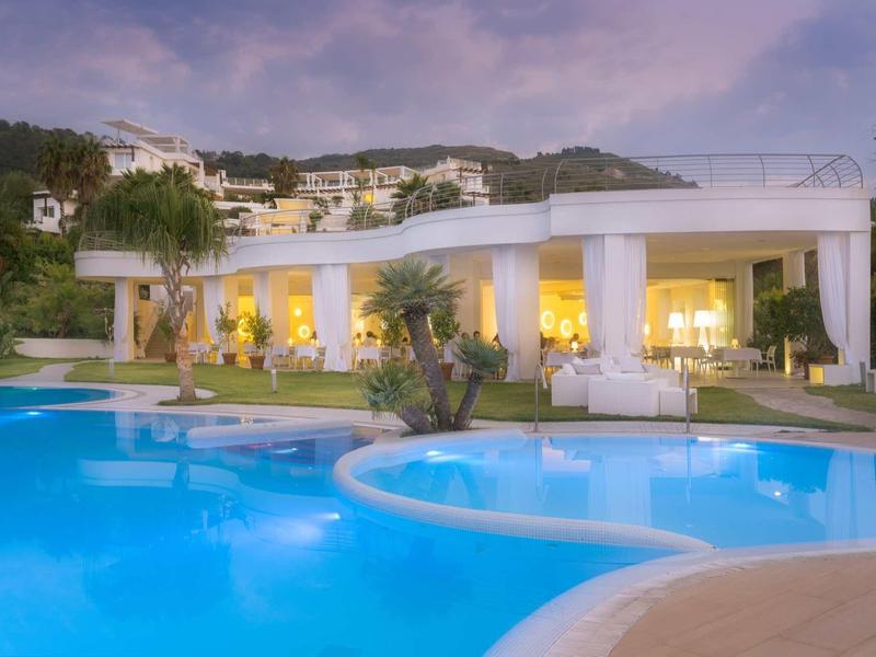 Wieczorny widok na oświetlony basen hotelowy z białym pawilonem i wzgórzami w tle.