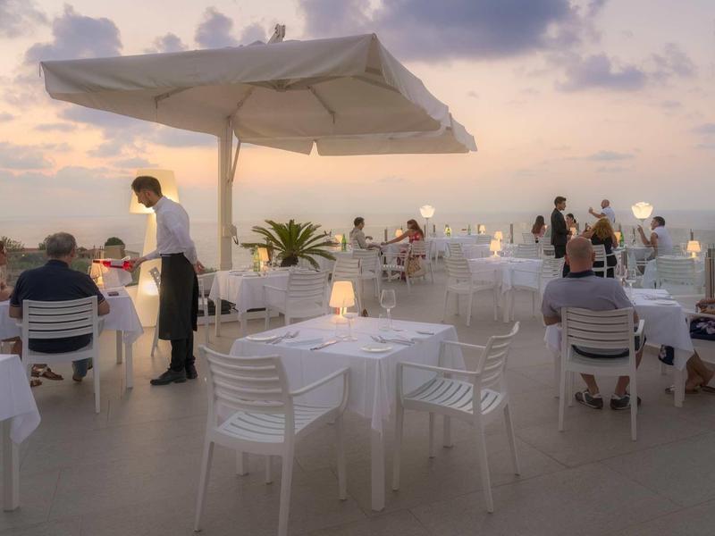 Taras na dachu z białymi stolikami i krzesłami, bufetem i gośćmi o zachodzie słońca.