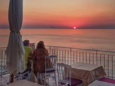 Osoba sedí na terase s stolky a židlemi a dívá se na západ slunce nad mořem.