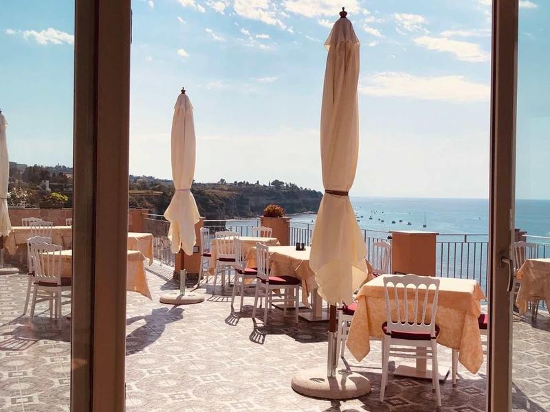 Terasa s prostřenými stoly a zavřenými slunečníky s výhledem na moře za jasné oblohy.