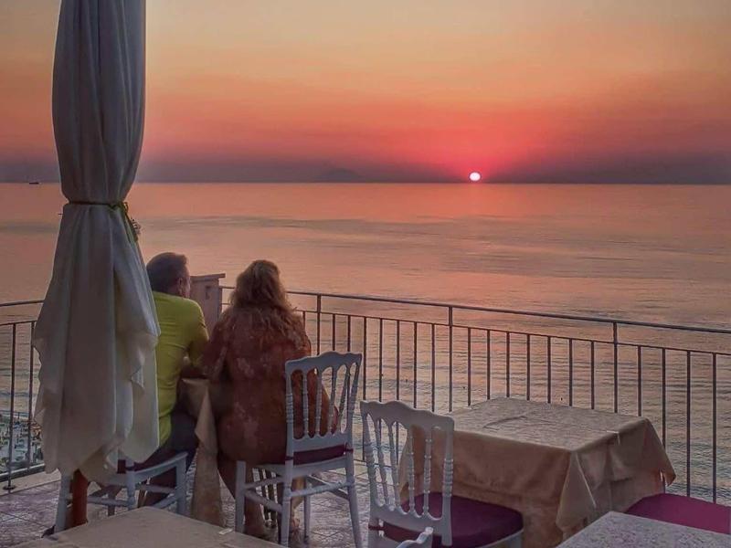 Osoba sedí na terase s stolky a židlemi a dívá se na západ slunce nad mořem.