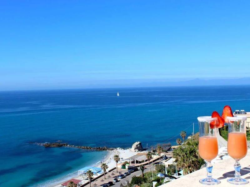 Pohled na modré moře s balkonem a dvěma nápoji v popředí.