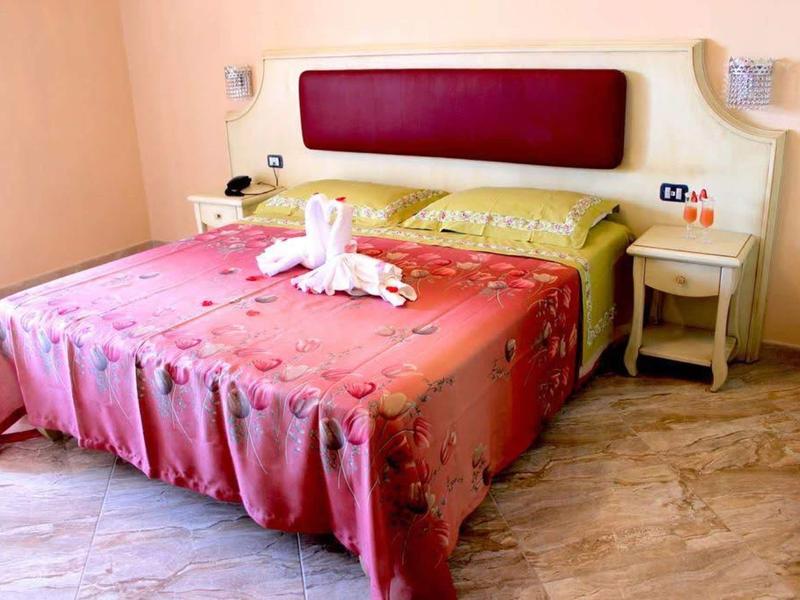 Pohodlná manželská postel s růžovým přehozem a labutí z ručníku v světlém hotelovém pokoji.