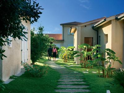 Weg mit Steinplatten führt durch grünen Garten zwischen hellen Häusern unter blauem Himmel.