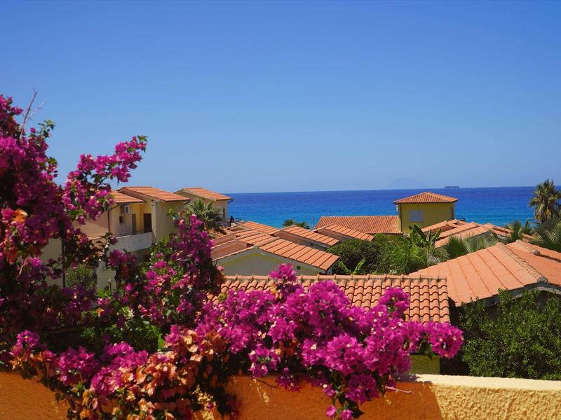 Blick auf rote Dachziegel, blühende Bougainvillea und blauen Himmel mit Meer im Hintergrund.