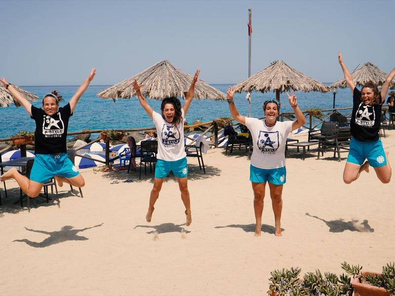 Vier Personen springen am Strand vor Sonnenschirmen, Meer und klarem Himmel, alle tragen schwarze Shirts.