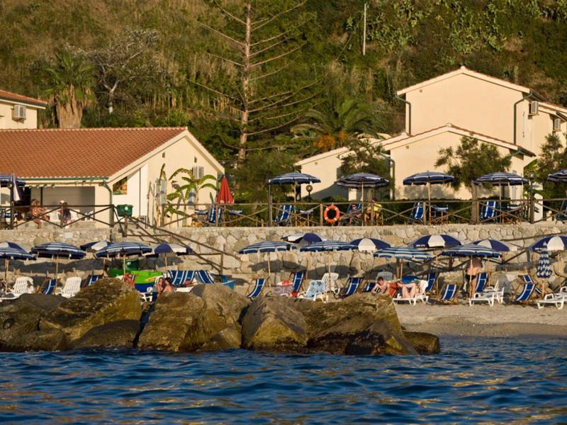 Strand mit blauen Sonnenschirmen, Felsen im Vordergrund und Häusern im Hintergrund bei Sonnenschein.