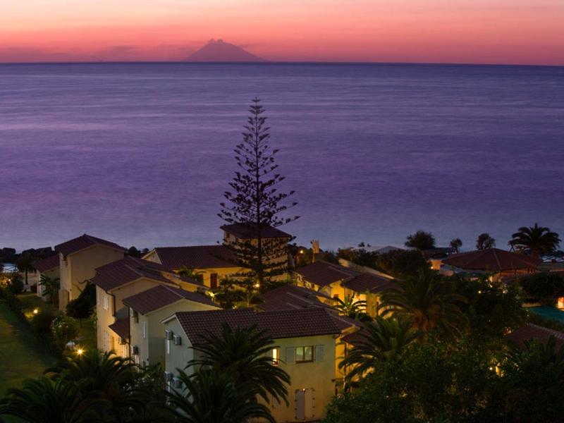Küstenlandschaft bei Sonnenuntergang mit Häusern, Palmen und ruhigem Meer im violetten Licht.