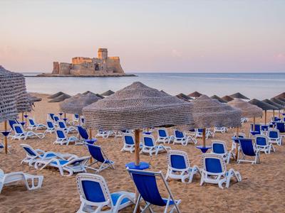 Spiaggia con sedie a sdraio blu vuote e ombrelloni, vista di un castello sul mare al tramonto.