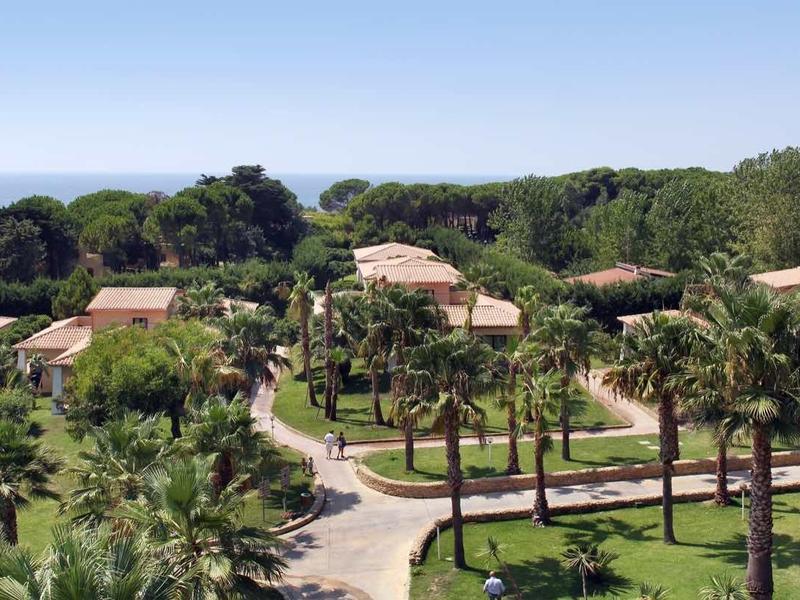 Blick auf ein sonniges Resort mit Palmen, Wegen und roten Dächern am blauen Meer im Hintergrund.