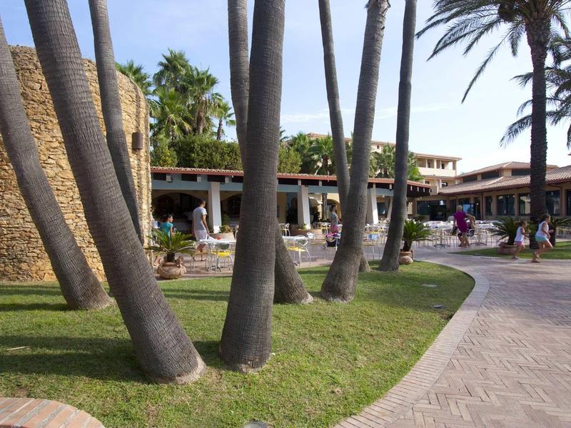 Palmen und grasbewachsene Flächen vor einem Gebäude mit Zinnenmauer und blauem Himmel.