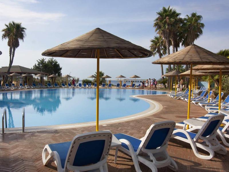Großer Pool mit blauer Wasseroberfläche, umgeben von Liegestühlen und Strohdach-Sonnenschirmen.