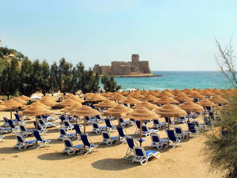 Strand mit vielen blauen Liegestühlen und Strohhütten, im Hintergrund Meer und eine Burg am Horizont.