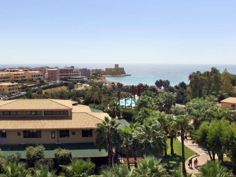 Blick auf Wohnhäuser mit roten Dächern, Palmen und blauem Meer unter klarem Himmel.