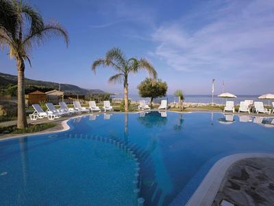 Blick auf einen Außenpool mit Palmen und Liegestühlen vor blauem Himmel.