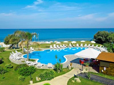 Buitenzwembad met ligstoelen en palmbomen aan zee onder een blauwe lucht.