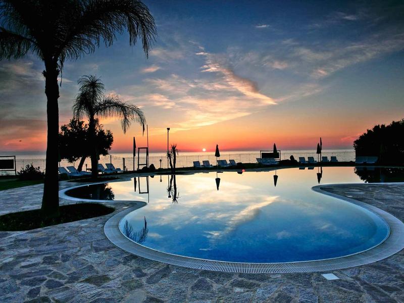 Zwembadgebied met palmbomen en zonsondergang achter de zee bij resort.