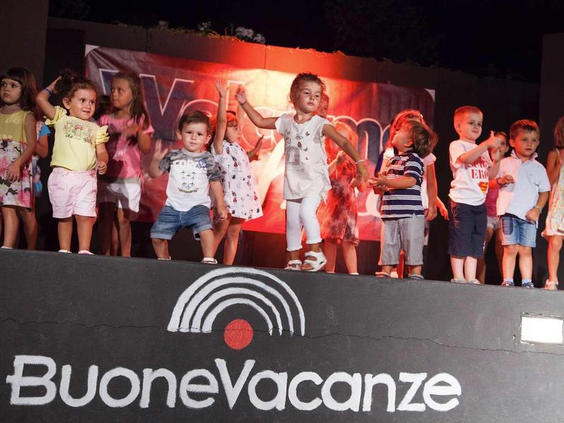 Kinderen dansen op een podium met het bord ‚Buone Vacanze‘ op de voorgrond.