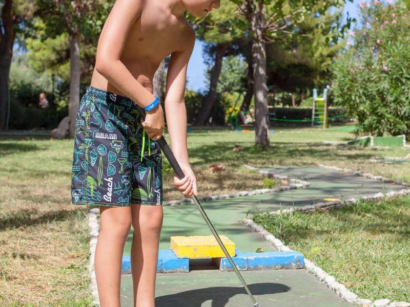 Een jongen speelt minigolven op een zonnige buitenbaan met bomen op de achtergrond.