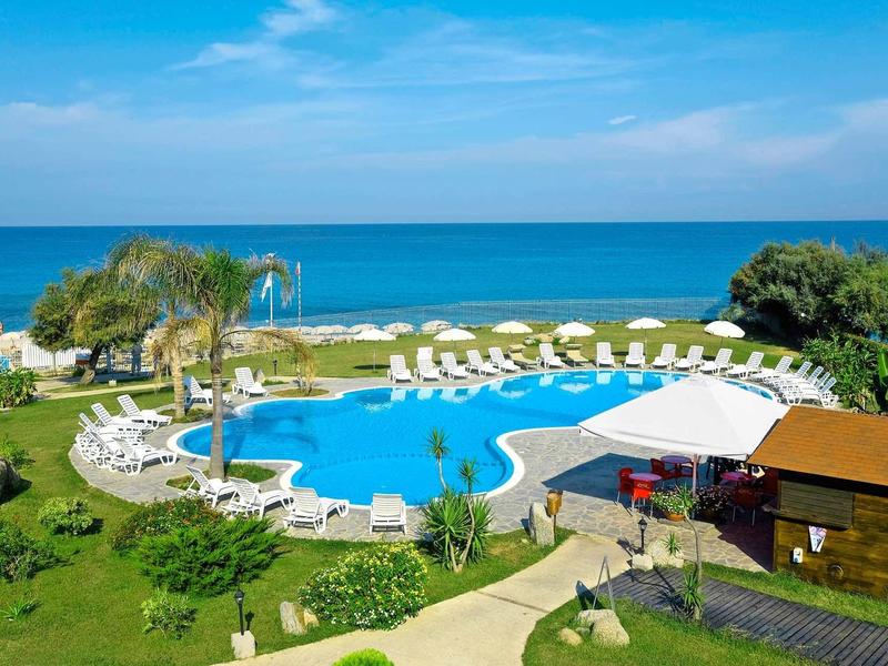 Buitenzwembad met ligstoelen en palmbomen aan zee onder een blauwe lucht.