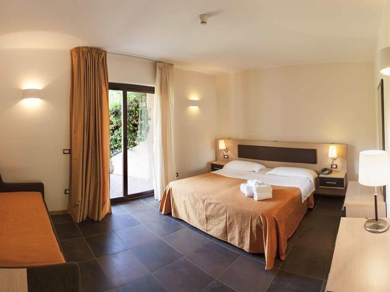 Lichte hotelkamer met tweepersoonsbed, bank, nachtkastjes en toegang tot het terras.