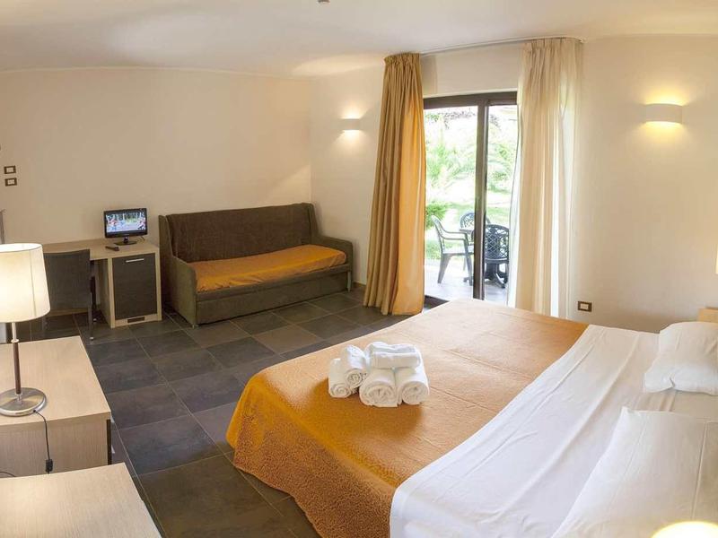 Hotelkamer met tweepersoonsbed, bank, bureau en balkon met uitzicht op groen.