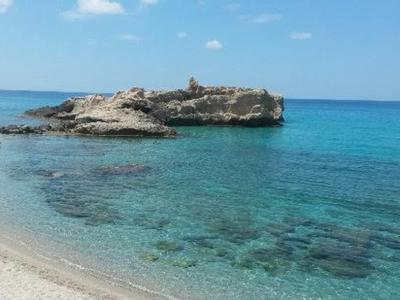 Čisté modré moře s písečnou pláží a skalnatým poloostrovem pod modrou oblohou.