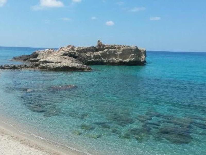 Čisté modré moře s písečnou pláží a skalnatým poloostrovem pod modrou oblohou.