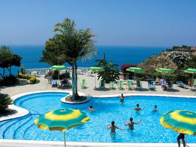 Swimmingpool mit Menschen, grünen Sonnenschirmen, Bäumen und Meer im Hintergrund bei klarem Himmel.