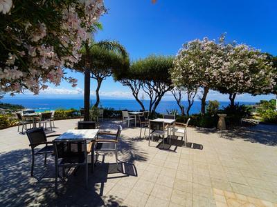 Terrasse mit Tischen und Stühlen unter blühenden Bäumen mit Meerblick bei klarem Himmel.