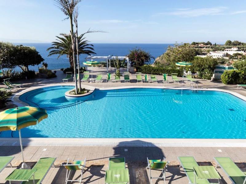 Zwembad met groene ligstoelen en uitzicht op de oceaan onder een blauwe lucht.