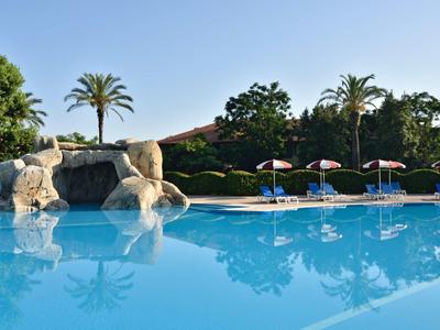Großer blauer Pool mit Wasserfall, Sonnenschirmen und Palmen im Hintergrund bei klarem Himmel.