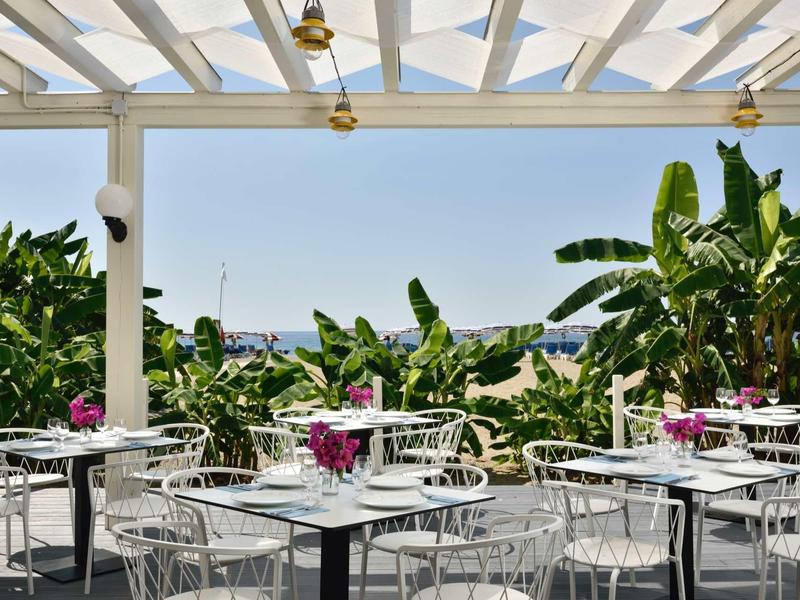 Terrazza coperta con tavoli e sedie bianche circondata da piante tropicali.