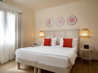 Licht hotelkamer met tweepersoonsbed, rode kussens en zachte verlichting.