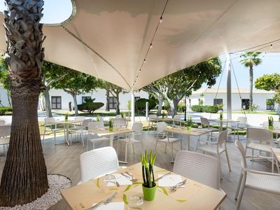 Moderne buitenruimte van een hotel met witte stoelen, tafels en zeilen onder bomen.