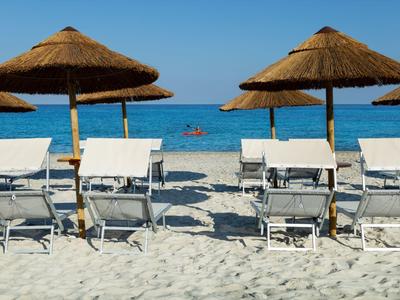 Strand met zand, meerdere witte ligbedden en strooien parasols bij de blauwe zee.