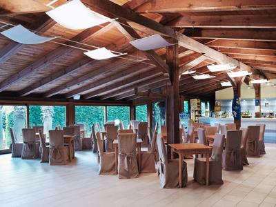 Rustykalna jadalnia z drewnianymi belkami, przykrytymi stołami i dużymi oknami z widokiem na zieleń.