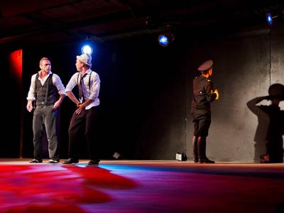 Scena teatralna z dwoma mężczyznami z przodu i osobą w mundurze w tle.