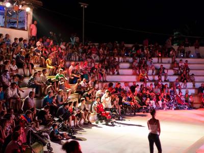 Duża grupa ludzi siedzi na arenie nocą, widok na scenę z osobą na pierwszym planie.