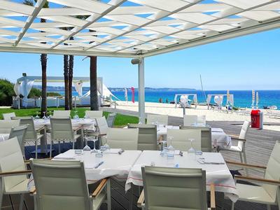 Taras restauracyjny z białymi stołami i krzesłami z widokiem na morze i plażę z parasolami.