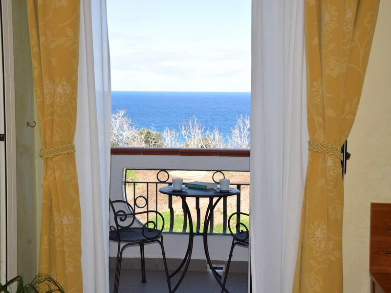 Balkon mit schwarzem Metalltisch und zwei Stühlen, vorne gelbe Vorhänge, Meer im Hintergrund sichtbar.